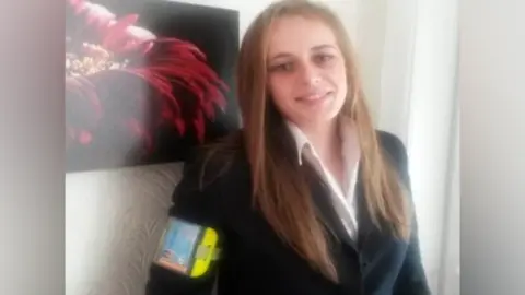 Chereiss Bailey is wearing a white shirt and black jacket and has long, light brown hair. She is leaning towards a wall and smiling at the camera. There is a picture on the wall behind her.
