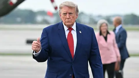 US Donald Trump looking serious, giving a thumbs up to the camera. He is wearing a blue suit with a red tie.