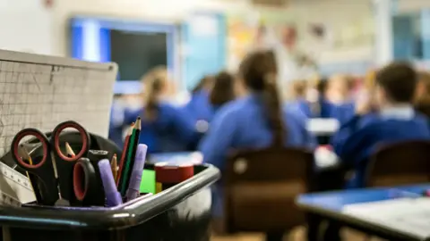 A close up of a stationary box filled with stationary in a classroom. The background is a blurred image of children sitting at their desks.