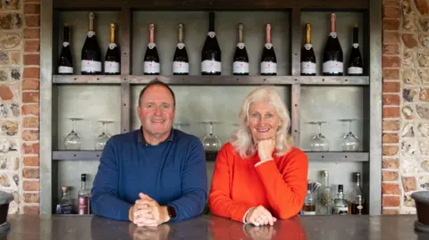 University of Brighton Mark and Sarah Driver are standing behind a counter with bottles of sparkling wine on the shelves behind them. Mark is wearing a blue top and Sarah is wearing red. The shelves are grey and set into a stone wall with a brick outline and the counter is shiny and grey.