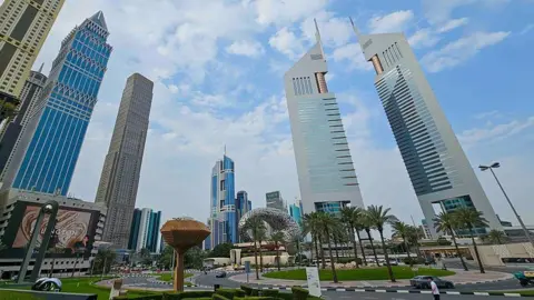 A view of the Dubai International Financial Centre, with palm trees, sculptures and skyscrapers visible in front of a blue sky