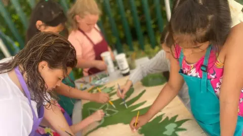 A group of young girls painting a large piece of artwork which is laid out on a table in front of them. It's a shared activity with them all working on the same piece. They hold small paintbrushes and wear colourful aprons.