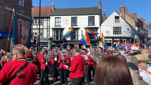A large brass band in red uniforms plays in the middle of street as crowds wave flags around them.