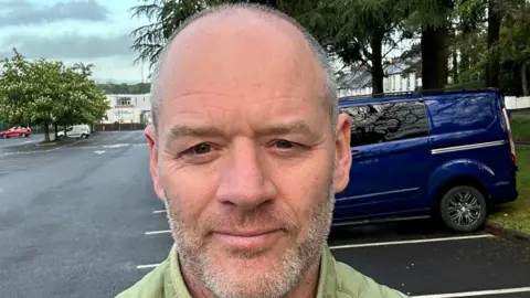 Timothy Shiels Tim has short grey hair, shaved tight to his head. His beard is a mix of grey and sandy brown. He is smiling softly. Behind him is a car park and some trees.