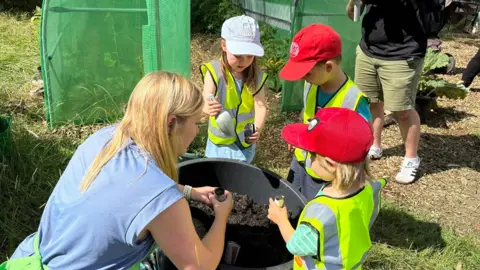 Martin Heath/BBC A woman with long blonde hair wearing a blue T-shirt is kneeling next to a bucket of soil with three children in yellow hi-vis vests. Two are wearing red caps and one has a white cap. There is a green growing tunnel behind them.