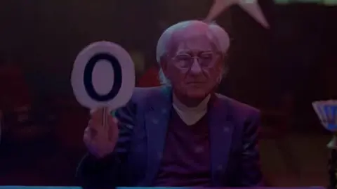 A man with long grey hair in a purple blazer and top holds up a padel with a 0 on it.