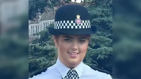 Surrey Police Hannah Byrne looks at the camera in her policing uniform. She is wearing a hat with the Surrey Police logo on it and a black and white checked neckerchief.