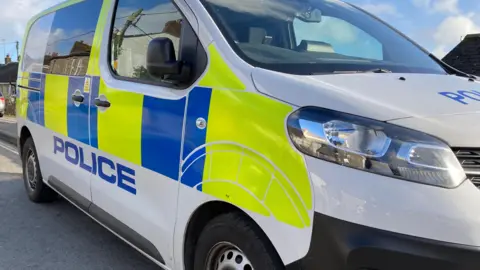 A picture of the side of a Devon and Cornwall police van. It is parked on a pavement on a long road 