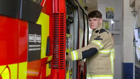 Mid and West Wales Fire and Rescue Service Bobi Jones smiles as he stands beside a fire engine in his firefighter's uniform. 