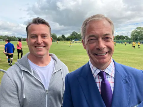 James McMurdock standing with Nigel Farage with a football match being played in the background