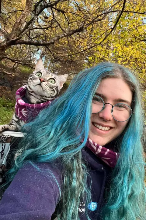 Lucy Francom A woman with dyed blue hair that is long and goes past her shoulders smiles at the camera. She is wearing glasses with blue frames and a purple fleece. She is carrying a grey cat with green eyes and wearing a purple harness. 