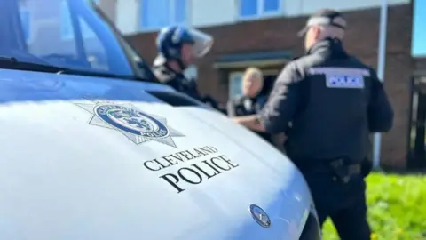 A police van in the foreground with the Cleveland Police logo on the bonnet. Three officers wearing dark clothing are talking to each other in the background.