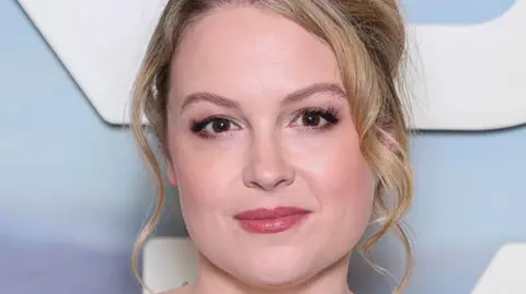 A woman with curly blonde hair in an up do. She is wearing a white dress and is looking at the camera with a neutral expression.