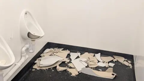 A white urinal in a public toilet has a large piece of porcelain missing and there is a pile of porcelain debris sitting on a dark floor beneath it