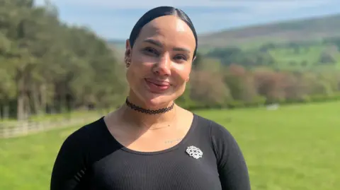 Georgia Levy-Collins/BBC A young woman with the Yorkshire moors in the background. She wears a black long-sleeved T-shirt and has slicked-back black hair. She also wears a black choker necklace.