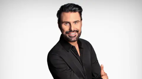 A man with dark brown hair smiling into the camera. He has a beard and white teeth. He is wearing a black shirt which is open at the top. 