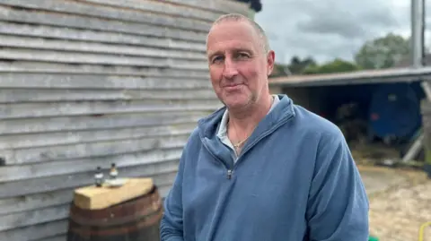 Tom Tame has very short light brown hair. He is wearing a blue zip jumper and is standing in front of a farm building