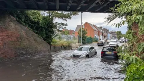 A silver Mercedes-type car is being driven into deep water, underneath a railway bridge. Another car, which is dark in colour, is being driven away from the camera through the water. There are terraced or semi-detached houses in the background.