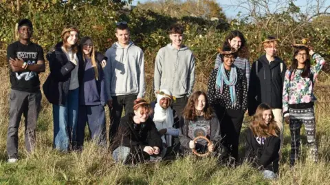 Connor Meadows A large group of young people pose for the camera at an Avon Wildlife Trust site in Begbrook in Bristol. Many of them are wearing hoodies and some are shielding their eyes as it is a sunny day. They are standing in a meadow with long grass and a hedgerow behind them