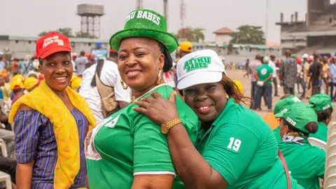 Grace Ekpu / BBC Two women in green T-shirts look at the camera smiling. On their heads are hats that say "Atiku Babes". They are outside in sports stadium.