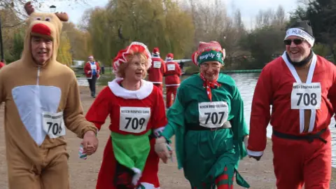 Four people dressed in festive attire, holding hands and running along Bedford's Embankment. The man on the left is wearing a reindeer onesie, a woman is wearing a Mrs Santa dress, an elf is next to her, and on the right is a man dressed in a Santa suit with a black hat. They all have numbers on them. Other runners in Santa suits, a river, a path and trees are behind them 