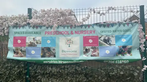 A view of a banner on a school gate. It shows details of Blossom preschool as well as that it has been rated as good by Ofsted. Blossom sits at the top of the fence.