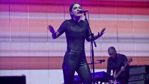 Sharon Van Etten performing onstage . She has short dark hair and is singing into the microphone while wearing all black. 