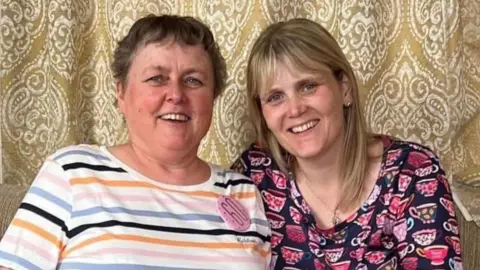 A photograph of a mother in her 60s and her daughter, who is in her 40s. They are sat in front of a gold curtain with a white pattern on it and the older woman, who has short dark hair, is wearing a colourful striped top with a 60th birthday badge pinned to it, while her daughter has shoulder-length blonde hair and is wearing a black top with pink mugs on it.