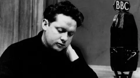 Dylan Thomas is seated in front of a BBC microphone in this black and white photo.