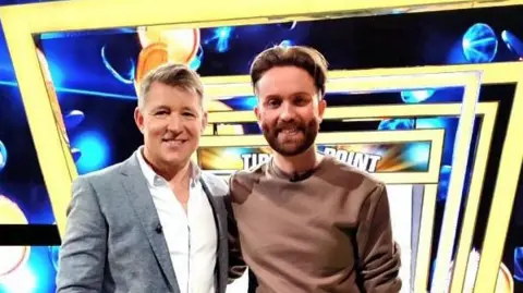 Publicity image of Ben Shephard and Jamie posing together on the Tipping Poin set, which has a blue background and a row of yellow square arches above.