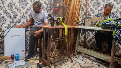 Two people in a room. A man on the left is looking at a power box. He is sitting down by a sewing machine and a fan. A woman is next to him, working at another sewing machine.