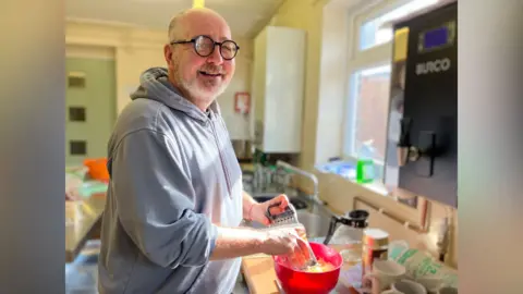 Jon Wright/BBC A smiling Andy Fell looks directly at the camera as he is grating cheese into a red bowl. He is stood at a counter in a kitchen, with sunlight streaming through a nearby window. Behind him is a kitchen sink and another workspace. He is wearing a grey hooded sweatshirt and black-rimmed glasses. 