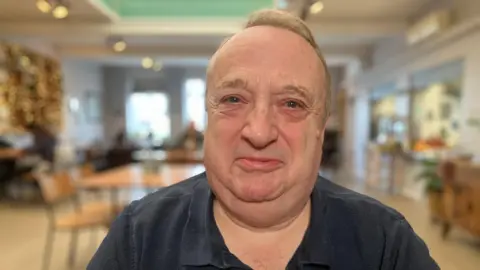 A middle-aged man with short grey hair, looking into the camera and smiling. he is wearing a black polo top and is sat in a cafe. Tables and chairs can be seen behind him. The background is blurred.