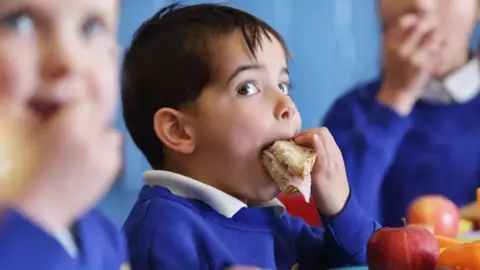 Getty Images A little boy with bark hair putting a sandwich in his mouth. He is wearing a blue jumper with with polo shirt underneath. Apples are in front of him and two other children are blurred beside him.