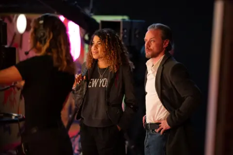 Studio Canal James McAvoy - a man with slicked back grey hair and a beard, wearing a black blazer, a white shirt and jeans - stands with his hands on his hips looking at a scene on a film set. Standing next to him is a young woman with dark curly hair, dressed in casual black clothes. In the foreground is a camera operator slightly out of focus.