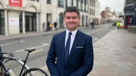 A man wearing a suit stands on a pavement outdoors. He is smiling at the camera. He is wearing a white shirt, navy tie and a navy blazer. A bike is parked next to him. People are walking on the pavement behind him. There is a road and buildings.