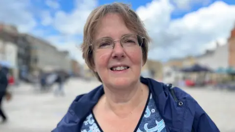 Emma Baugh/BBC A woman with short blonde hair wearing a blue jacket and a patterned top standing in a street with shoppers in the background.