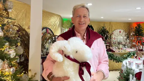 A man wearing a pink shirt with a red jumper draped over his shoulders. He is holding a large sheep teddy. He is standing inside a shop which has Christmas items on display. Decorated Christmas trees are also on display. A gold fabric is draped on the walls inside the shop.