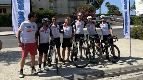 Seven men who have finished a cycling challenge from Leeds to Nice, they are standing a row with their arms around each other, some with their bikes, some wearing helmets, all smiling.