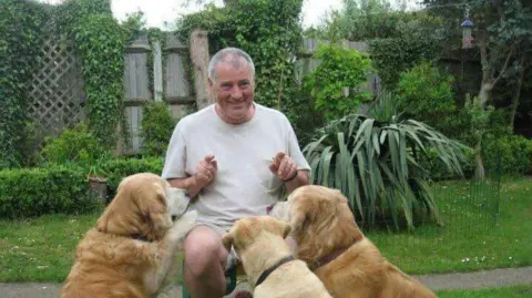 Anne McEntee A man in a tidy, green garden sat on a stool. Three golden dogs and jumping up onto his lap. He is smiling. 