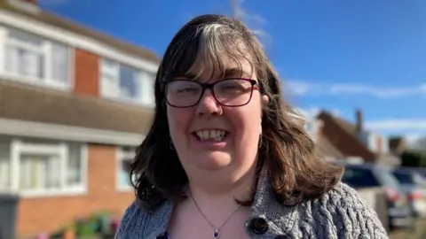 A middle aged woman who has a brown bob with grey roots smiles at the camera. She is wearing black glasses, a silver necklace with a black heart stone and a knitted grey cardigan. It is a sunny day and she is stood in front of her house.