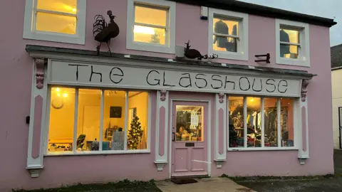 The Glasshouse building in Wigton. The two-storey building is painted pink with white details around the windows. Inside the seating of the cafe is visible, as well as the thrift shop.