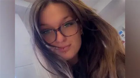 Alysha Ridley A selfie of a woman with long brown hair and brown glasses. She has a piercing through her bottom lip