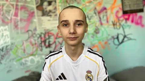 Leo sits on his bed in his bedroom, behind him is a colourful graffiti wall. He is wearing a signed Real Madrid top which was gifted to him by Jude Bellingham. He has a buzz cut.