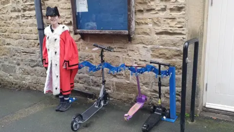 Leeds City Council A child dressed in a red ceremonial robe with white trim and a black tricorn hat is standing next to a wall‑mounted scooter rack. Three scooters are lined up in the rack behind them, with a blue noticeboard and a white door in the background.