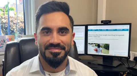 Dr Ali Ilyas, a GP from Cleethorpes, looks directly into the camera wearing a white shirt and sat in front of his computer in his GP surgery.