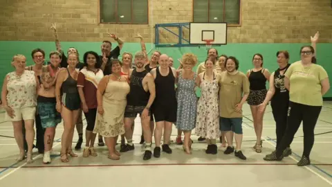 Contributed A group of people standing looking at the camera smiling. Some have their arms up and are waving or pointing. They are standing in a gymnasium with a teal wall behind them and a basketball hoop on the wall. 
