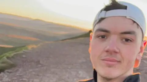 Jordan Harries A man smiling a the camera stood on top of a mountain with the sun rising in the background.