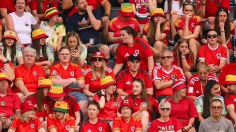 FAW A picture of a crowd of fans sitting during the match. They are all wearing red shirts. Some have their arms folded while others look disapointed by the result.