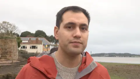 David Reed MP is wearing a beige crewneck sweater and a burnt orange and grey waterproof jacket and is standing on the shore at Lympstone.